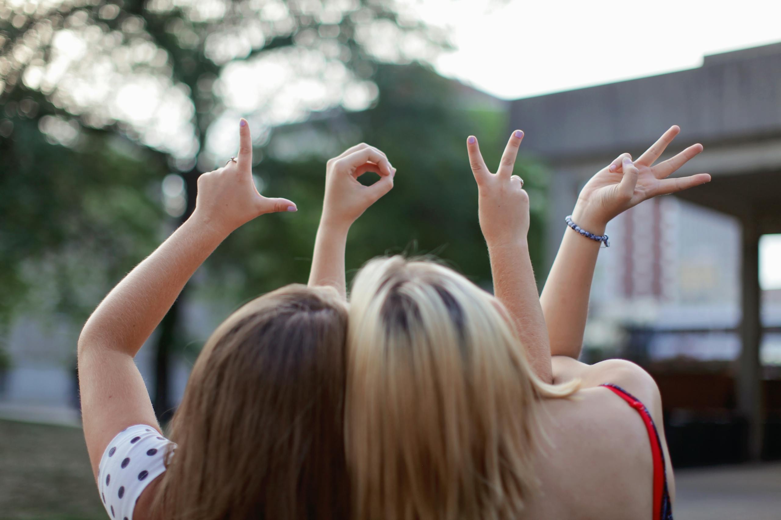 Two young women forming the word 'LOVE' with hand signs, symbolizing friendship in a summer setting.